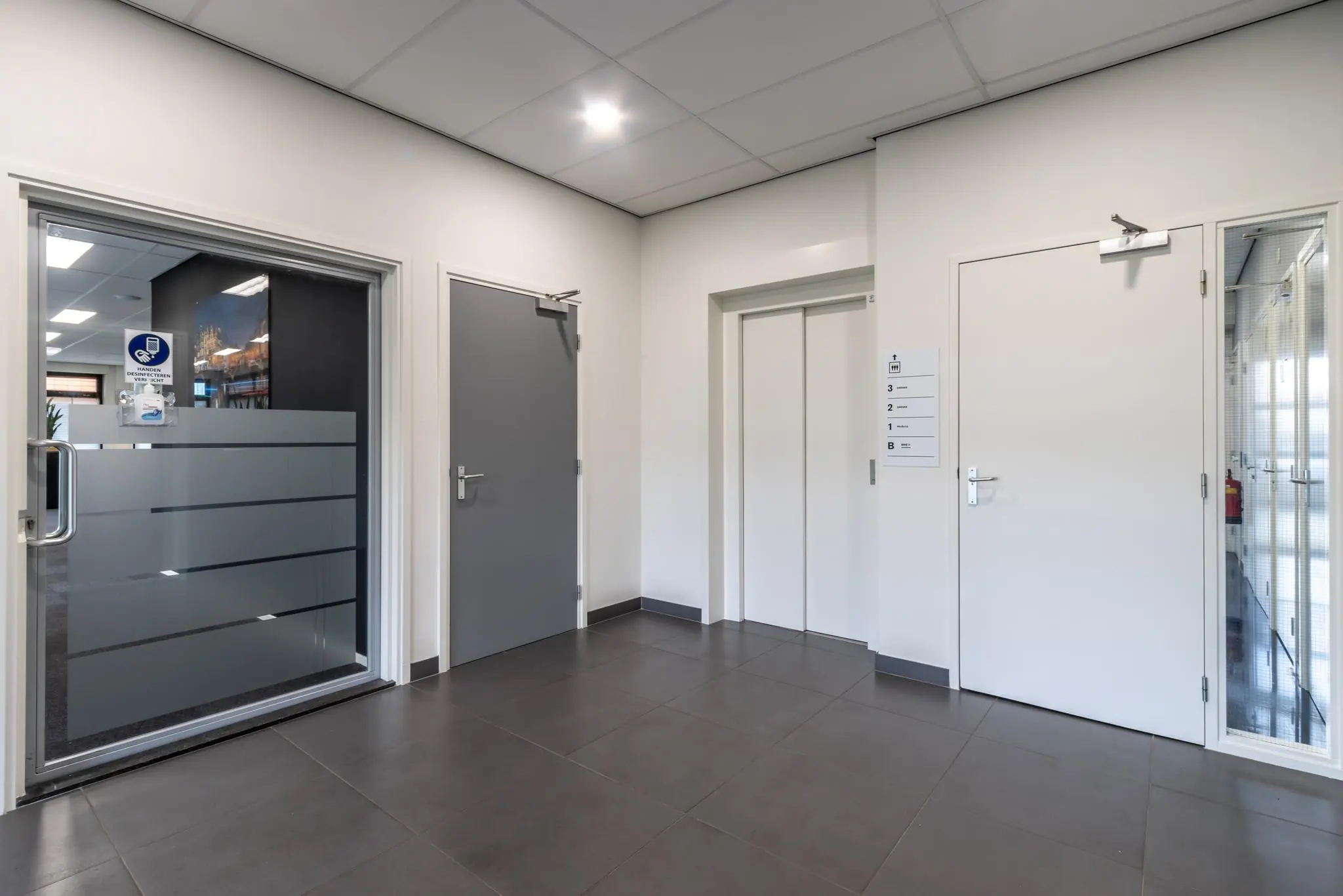 Moderne kantoorhal met een lift, grijze vloertegels en meerdere gesloten deuren aan de Lage Biezenweg.